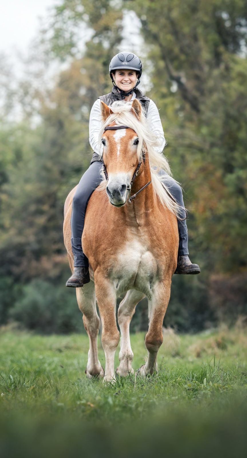 Reiterin reitet ihren Haflinger entspannt über eine Wiese und kommt lächelnd auf die Kamera zu
